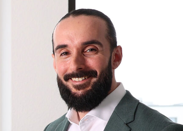 Mann mit dunklem Haar und Bart, lächelnd, trägt ein weißes Hemd und eine grüne Jacke.