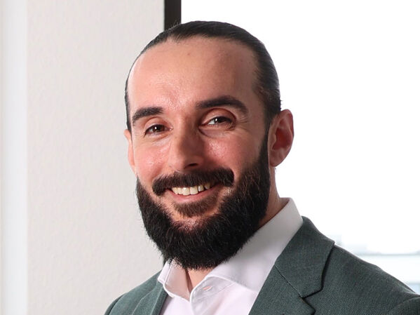 Mann mit dunklem Haar und Bart, lächelnd, trägt ein weißes Hemd und eine grüne Jacke.